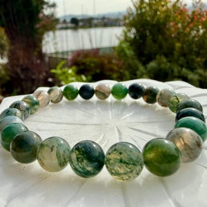 May include: A circular beaded bracelet composed of polished moss agate stones. The beads feature varying shades of translucent green, deep forest green, and milky white, with intricate dark green dendritic inclusions visible within each sphere.
