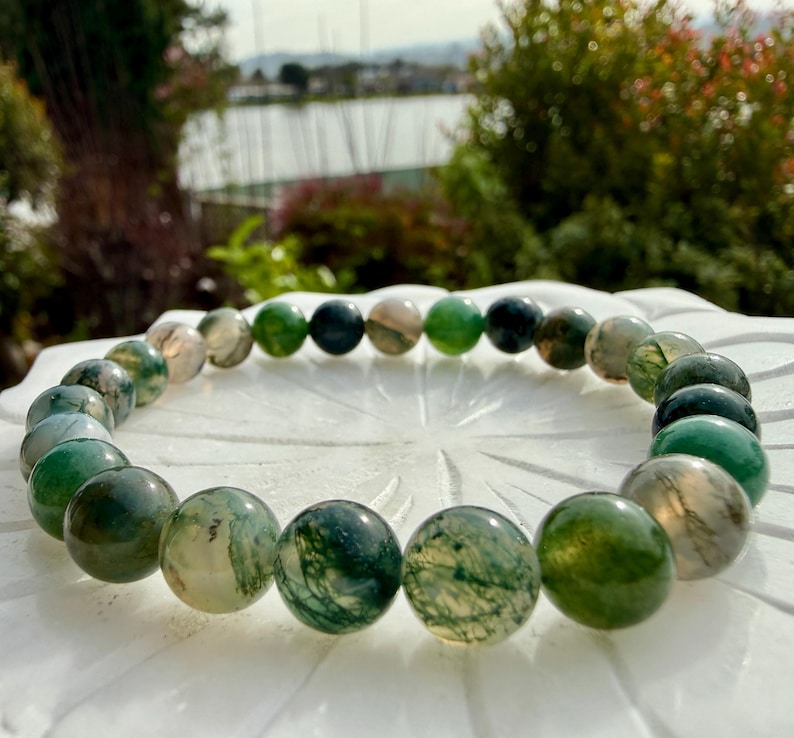 May include: A circular beaded bracelet composed of polished moss agate stones. The beads feature varying shades of translucent green, deep forest green, and milky white, with intricate dark green dendritic inclusions visible within each sphere.