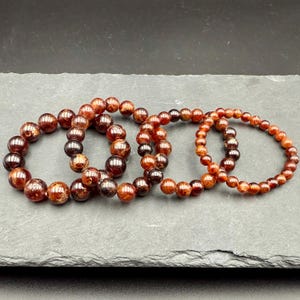 Pode incluir: Tr&ecirc;s pulseiras circulares feitas de contas de granada vermelho-profundo polidas. As pulseiras variam no tamanho das contas, com as maiores &agrave; esquerda e as menores &agrave; direita, todas com uma superf&iacute;cie brilhante e varia&ccedil;&otilde;es de cor naturais.