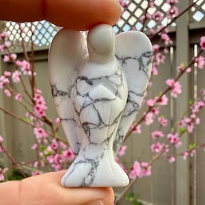Pode incluir: Pequena estatueta de anjo branco com veios cinzentos. O anjo tem as asas abertas e é segurado entre os dedos. O fundo apresenta flores cor-de-rosa e uma cerca de madeira.