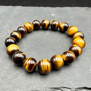 May include: A beaded bracelet featuring polished tiger eye stones. The bracelet is made of round, brown and gold beads with a striped pattern. The beads are strung together to form a circular shape.