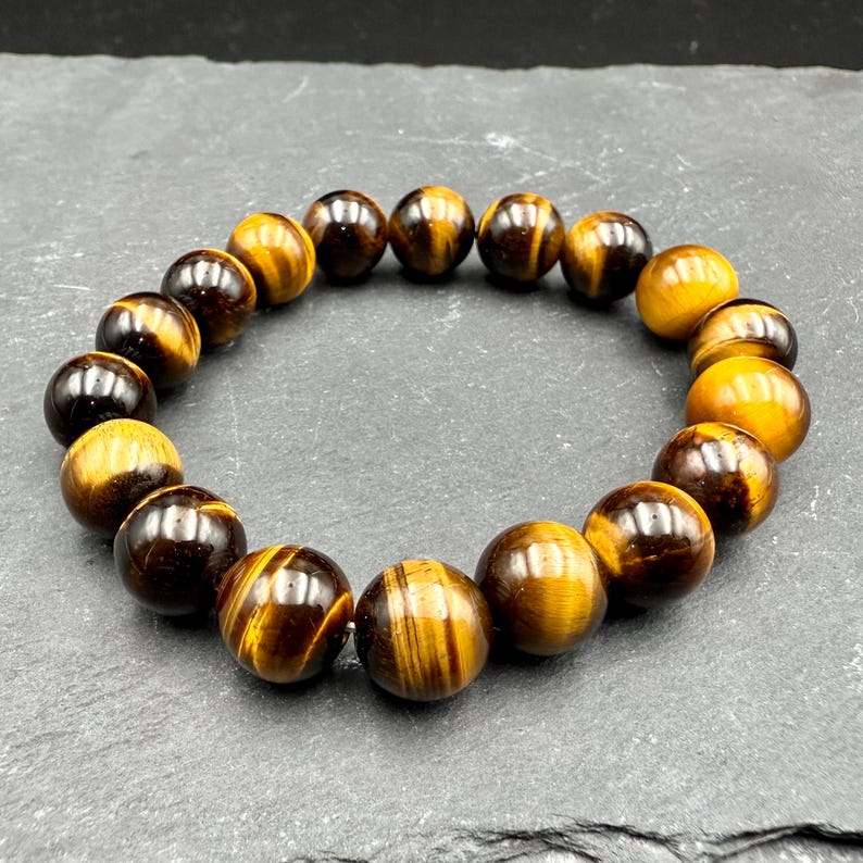 May include: A beaded bracelet featuring polished tiger eye stones. The bracelet is made of round, brown and gold beads with a striped pattern. The beads are strung together to form a circular shape.