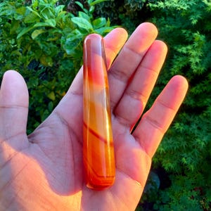 Peut inclure: Une pierre polie et allongée aux tons orange, rouge et jaune. La pierre a une finition lisse et brillante et semble être un matériau naturel. La forme est cylindrique avec des extrémités arrondies. La pierre est tenue dans une main sur fond de feuillage vert.