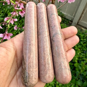 Op de afbeelding: Drie roze en grijze stenen massage staven. Elke staaf is cilindrisch met een afgeronde punt. De staven worden in een hand gehouden, met een achtergrond van groen gebladerte en roze bloemen.