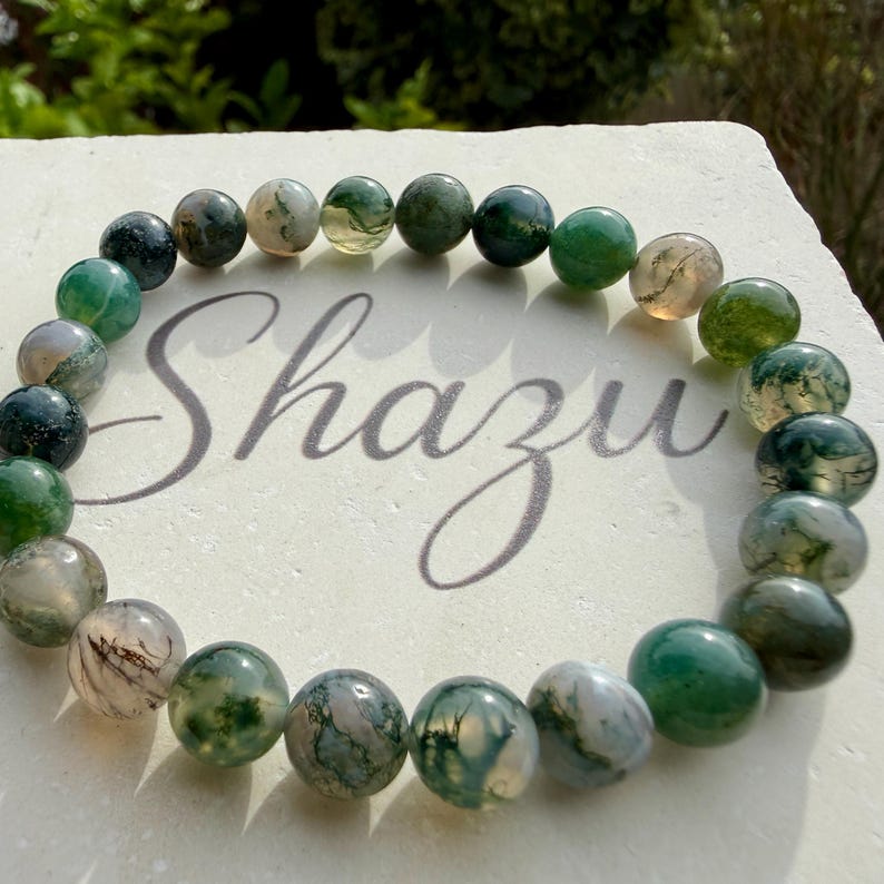 May include: A circular bracelet composed of polished, spherical moss agate beads in varying shades of translucent green, dark forest green, and milky white with natural inclusions, arranged in a continuous loop on a textured surface.