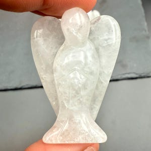 May include: A clear quartz angel figurine with outstretched wings. The angel is carved with detailed features, including a flowing gown and defined facial features. The crystal is translucent, allowing light to pass through, and is held by a person's fingers.
