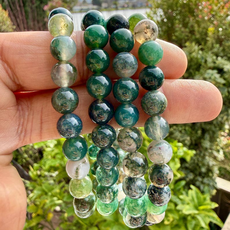 May include: Three strands of polished moss agate beads featuring a variety of green, white, and translucent tones. Each bead displays unique natural inclusions and patterns, ranging from deep forest green to pale mint and cloudy white hues.