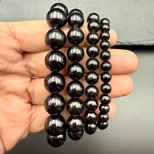 May include: Several black beaded bracelets of varying sizes are displayed. The beads are round and have a glossy finish. The bracelets are held in a hand, showcasing the different sizes and styles of the jewelry.