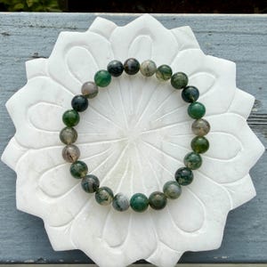 May include: A circular beaded bracelet featuring smooth, polished round beads in varying shades of forest green, moss green, and translucent grey with natural inclusions, arranged in a continuous loop on a white, flower-shaped stone base.