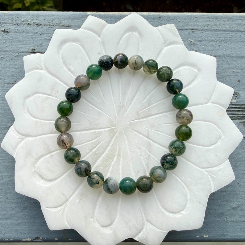 May include: A circular beaded bracelet featuring smooth, polished round beads in varying shades of forest green, moss green, and translucent grey with natural inclusions, arranged in a continuous loop on a white, flower-shaped stone base.