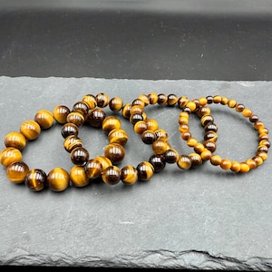 May include: Four beaded bracelets made of tiger's eye gemstones. The bracelets are arranged on a dark gray surface. The beads are a mix of brown, gold, and black, with a polished, round shape. The bracelets vary slightly in bead size.