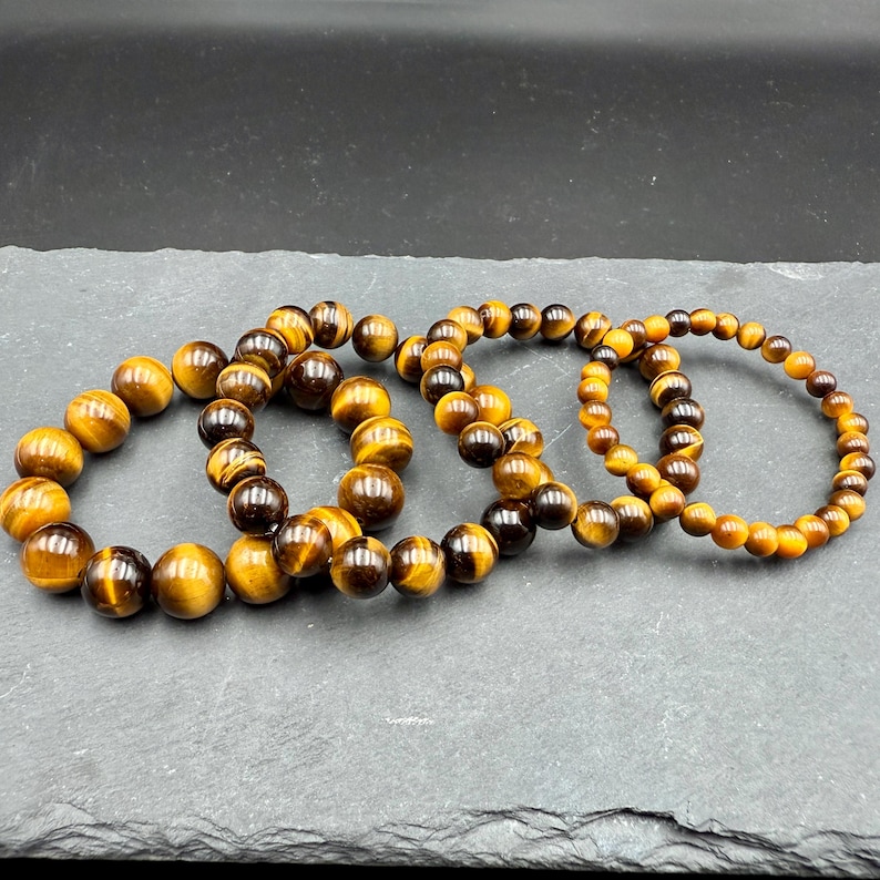 May include: Four beaded bracelets made of tiger's eye gemstones. The bracelets are arranged on a dark gray surface. The beads are a mix of brown, gold, and black, with a polished, round shape. The bracelets vary slightly in bead size.
