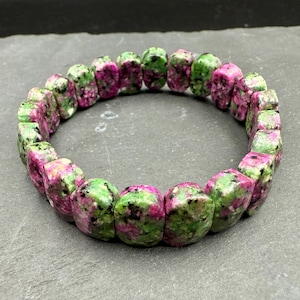 May include: A bracelet composed of rectangular ruby zoisite beads. The beads are a mix of green and magenta hues, with a mottled pattern. The bracelet is formed in a circular shape, showcasing the natural stone's colours and textures.