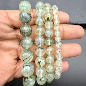 May include: Three beaded bracelets made of polished, translucent green rutilated quartz spheres. The beads vary in size across the three strands, featuring internal dark needle-like inclusions and cloudy mineral patterns within the smooth stone surfaces.