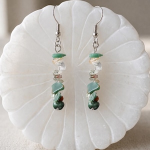May include: A pair of dangle earrings featuring silver-toned metal hooks and a vertical arrangement of small, irregular, tumbled gemstone chips in shades of green, clear, and brown, suspended in a linear design from the top metal loop.