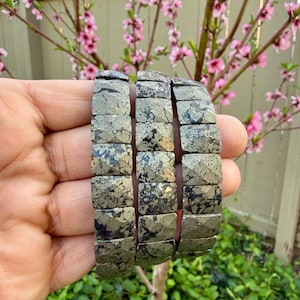 May include: Three pyrite bracelets with a faceted design. The bracelets are a mix of metallic gold and dark gray, with a textured appearance. The bracelets are shown against a backdrop of pink blossoms and green foliage.