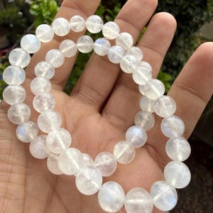 May include: Two strands of white, round gemstone beads. The beads have a translucent quality with subtle iridescent flashes. The bracelet is held in a hand, with a blurred green background.