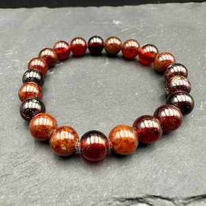 Puede incluir: Un brazalete de cuentas compuesto por piedras preciosas redondas y pulidas de color marrón rojizo. Las cuentas varían en color, algunas aparecen más oscuras y otras más claras, creando un degradado natural. El brazalete está ensartado en una banda elástica.