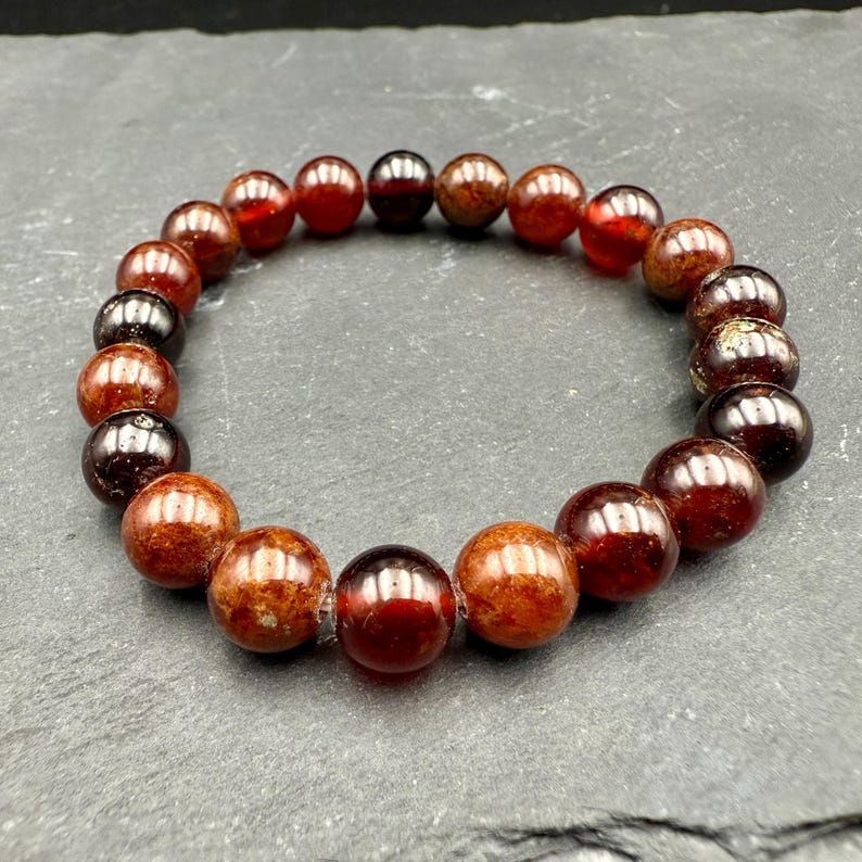 Pode incluir: Uma pulseira circular composta por contas esf&eacute;ricas polidas em tons variados de vermelho profundo, bord&ocirc; escuro e laranja acastanhado. As contas est&atilde;o enfiadas num la&ccedil;o cont&iacute;nuo, exibindo uma superf&iacute;cie lisa e reflexiva com varia&ccedil;&otilde;es.