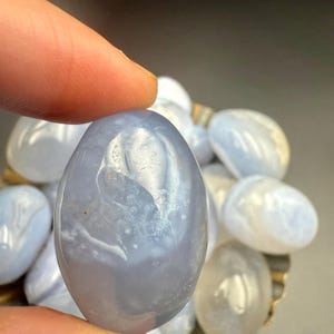 Op de afbeelding: Een gladde, ovaalvormige blauwe kantagaat edelsteen wordt tussen vingers gehouden. De steen heeft een doorschijnend uiterlijk met witte en lichtblauwe patronen. Andere soortgelijke stenen zijn zichtbaar op de achtergrond.