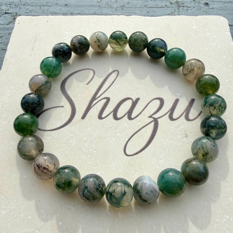 May include: A circular bracelet composed of polished, spherical moss agate beads in varying shades of deep forest green, translucent white, and earthy brown. The beads are strung together in a continuous loop, displaying natural inclusions.