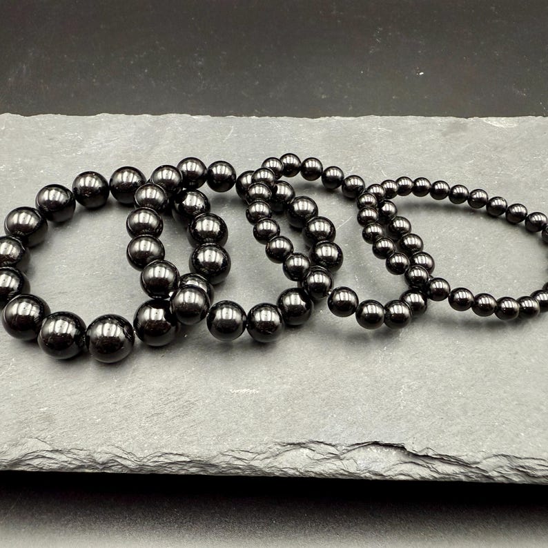 May include: Four black beaded bracelets of varying sizes are displayed on a gray stone surface. The bracelets are made of smooth, round beads with a glossy finish. The beads range in size, creating a layered effect.