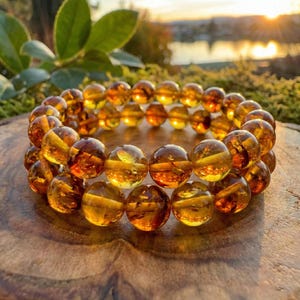 May include: A double-strand bracelet composed of amber-toned, spherical beads. The beads are translucent, displaying hues from pale yellow to rich brown. The bracelet is presented on a wooden surface, with natural light enhancing the beads' appearance.