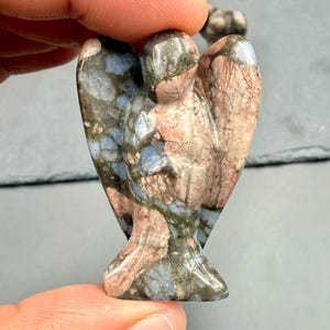 Könnte beinhalten: Eine geschnitzte Steinengelfigur mit ausgebreiteten Flügeln. Der Engel ist hauptsächlich in Blau-, Braun- und Grautönen gehalten und hat eine strukturierte Oberfläche. Die Flügel sind weit ausgebreitet und die Figur hat einen ruhigen Ausdruck. Ein kleiner Dekorationsartikel.
