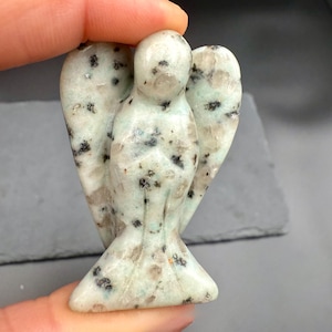 May include: A carved stone angel figurine in shades of pale green, white, and gray, speckled with black. The angel has outstretched wings and a flowing gown. The figurine is held by a person's fingers.