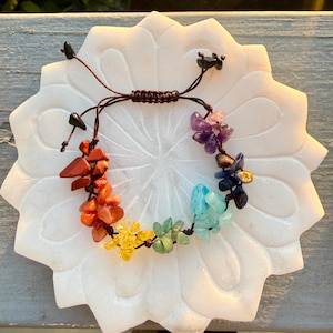 Puede incluir: Un brazalete de chakras con piedras preciosas de colores: rojo, naranja, amarillo, verde, azul, índigo y violeta. Las piedras están ensartadas en un cordón marrón. El brazalete se exhibe en un plato blanco con forma de flor.
