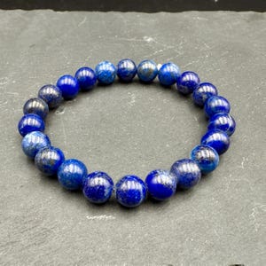 May include: A stretch bracelet composed of round, polished lapis lazuli beads. The beads feature a deep royal blue color with subtle, natural dark and gold-toned inclusions throughout the stone surfaces, arranged in a circular loop pattern.