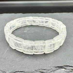 May include: A clear quartz bracelet with a rectangular, segmented design. The bracelet is made of translucent, polished quartz stones, and is displayed on a dark gray surface. The bracelet is a piece of jewelry.