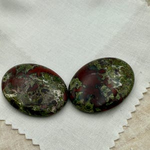 May include: Two oval-shaped stones with a mix of deep red and green hues. The stones have a polished appearance and are set against a white fabric background. The stones are likely used for decorative or spiritual purposes.