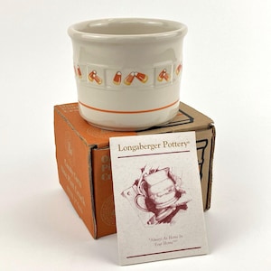 May include: A cream-colored ceramic crock with a candy corn pattern around the rim and an orange stripe near the base. The crock sits on a brown cardboard box labeled Candy Corn One Pint Crock, accompanied by a Longaberger Pottery product card.
