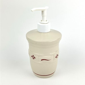 May include: Cream-colored ceramic soap dispenser with a white pump. The dispenser has a cylindrical shape with a slightly rounded top and is decorated with red floral accents and a horizontal line near the middle.
