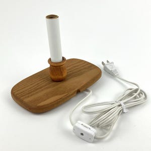 May include: A wooden lamp base with a rectangular, rounded-edge platform and a central vertical socket. The fixture includes a white electrical cord with a plug and an inline switch. The wood features a visible grain pattern and a light brown finish.
