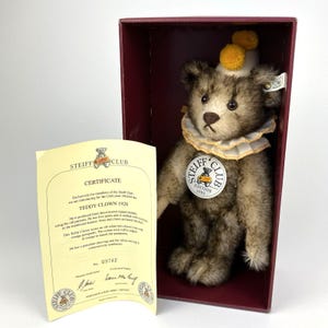 May include: A Steiff Club Teddy Clown 1928 plush bear in a burgundy box. The bear has brown and tan fur, a white ruffled collar, and a small hat with yellow pom-poms. A certificate is visible.