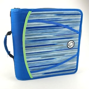 May include: A blue zippered binder featuring a horizontal motion-blur pattern in shades of blue, white, and gray. The binder includes a bright lime green accent strip along the left side, a black fabric handle, and a circular logo on the front cover.