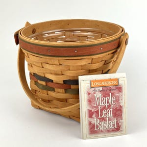 May include: A round woven wooden basket with a swing handle, featuring a horizontal band in rust and green. The basket includes a clear plastic liner and a product card labeled Longaberger 1996 Shades of Autumn Maple Leaf Basket.