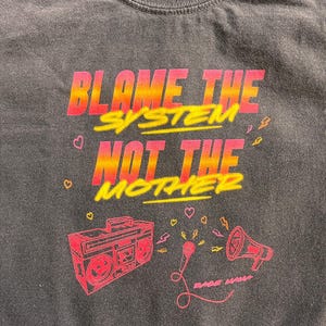 Puede incluir: Camiseta gris oscuro con el texto "BLAME THE SYSTEM NOT THE MOTHER" en rosa y amarillo. La camiseta presenta un diseño rojo de una radio, un micrófono y un megáfono. También se incluyen pequeños corazones y rayos.