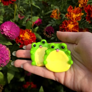 May include: Two small, bright green frog-shaped trinket dishes with yellow bellies, black eyes, and tiny pink tongues sticking out. These smooth, glossy ceramic-style figures feature raised arms and a flat, rounded base for holding small items.