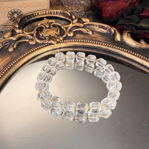 May include: A clear, faceted crystal bracelet with gold-colored spacers. The bracelet is displayed on a reflective surface within a decorative, ornate gold-framed mirror. The background includes crystals and a dark red fabric.