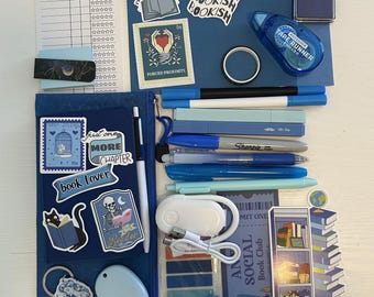 May include: A collection of blue-themed stationery and reading accessories including a reading log, a pencil case with stickers, various pens, a tape runner, a charging cable, bookmarks, a small stamp, and a blue bow clip arranged on a surface.
