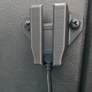 Puede incluir: Un soporte de cable USB de plástico negro montado con dos tornillos. El soporte tiene forma rectangular con dos soportes verticales para sujetar el cable. El cable es negro y está conectado a un dispositivo.