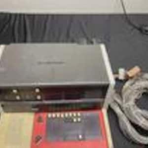 May include: A black and silver electronic device with a series of buttons and a display screen. The device is rectangular and has a white border. A red control panel and a bundle of gray cables are also visible.