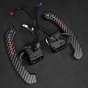Op de afbeelding: Twee carbon fiber schakelflippers met rode en blauwe LED-verlichting. De shifters hebben een zwarte basis en zijn verbonden door draden met rode, blauwe en groene connectoren. De achtergrond is een donkergrijze ondergrond.