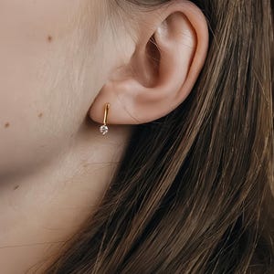 May include: A close-up of a gold earring with a small, clear gemstone. The earring is a simple, elegant design, with a small bar that curves slightly. The earring is in the ear of a person with long, brown hair.