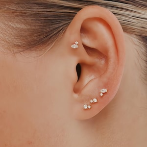 May include: Close-up of an ear adorned with multiple small, gold-toned earrings. The earrings feature clear, sparkling gemstones in various shapes, including round and marquise cuts. The jewelry is delicate and adds a touch of elegance.