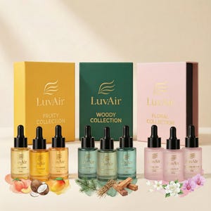 Op de afbeelding: Een productfoto toont drie sets LuvAir etherische olieflessen met dozen. De Fruity, Woody en Floral collecties worden gepresenteerd met hun flessen. Elke fles heeft een zwarte druppelaar. Decoratieve elementen zoals fruit en bloemen zijn ook inbegrepen.