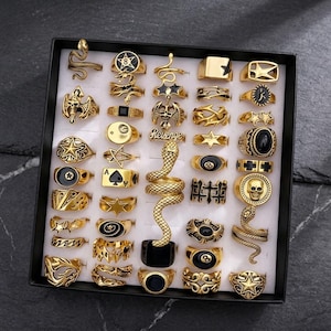 May include: A collection of gold-toned rings arranged in a black display box. The set features various designs including snakes, skulls, stars, playing card suits, and billiard balls. Some rings incorporate black enamel accents and the word Revenge.